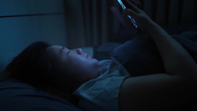 Asian woman unable to sleep, using her smartphone before bedtime and enjoying short-form video content on a platform, with the phone�s light illuminating her face
