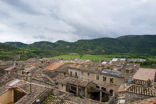 Traditional stone buildings with weathered terracotta rooftops overlook lush fields and forested hills in Ainsa, under a cloudy sky. The rustic textures and muted tones evoke a tranquil rural