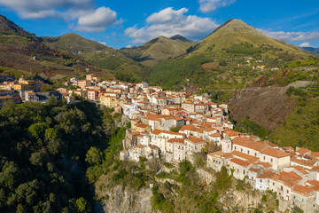 Fototapeta premium Drone view of Tortora, Italy, with clustered stone houses and terracotta roofs stretching along a rugged ridge, surrounded by green wooded hills and distant mountains under a bright blue sky.