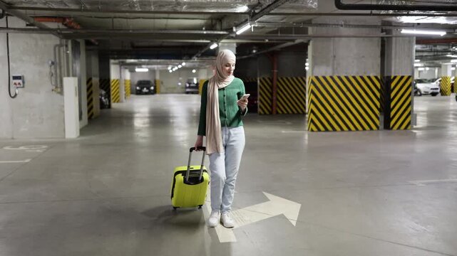 A woman wearing a hijab walks through a parking garage while looking at her phone and pulling a suitcase