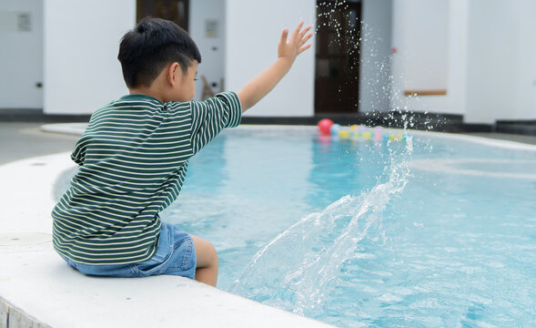 Little happy boy sitting and splashing on side of swimming pool playing by his feet in water