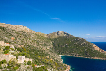 Obraz premium Aerial view of a tranquil bay framed by rugged hills and traditional stone buildings nestled among greenery on the Mani peninsula, Greece, under a vivid blue sky.