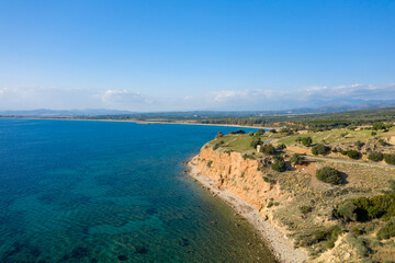 Obraz premium Aerial view of a rugged coastline with orange cliffs meeting transparent blue water, surrounded by open countryside and distant mountains beneath a cloud-streaked sky. Sunlight enhances the vivid
