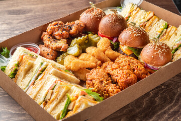 A tempting assortment of party foods in a cardboard platter box, featuring mini cheeseburger...