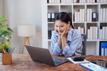 Fototapeta premium Asian woman smiling, working on her laptop at a wooden desk in an office or library environment, engaged in online communication or learning, representing remote work and student life