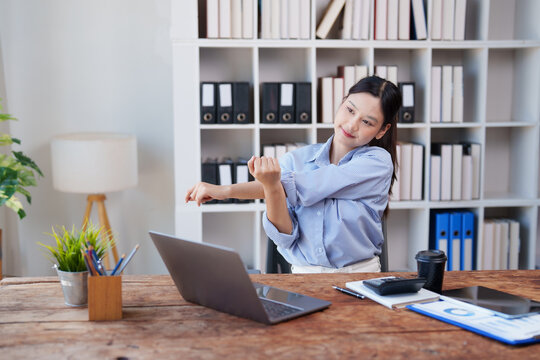 Young businesswoman stretching her arms and shoulders while working on laptop at a wooden desk in a modern office, taking a necessary break for improved posture and wellness