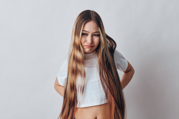 Young girl with long straight hair wearing white crop top stands against plain background with hands on hips looking at camera in confident casual pose for portrait. © SHOTPRIME STUDIO