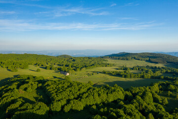 Fototapeta premium A solitary stone house nestles among rolling green fields and scattered trees, surrounded by expansive hills and forests under a bright blue sky in the Ardeche countryside.