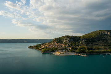Fototapeta premium Aerial view of Bauduen village bathed in soft golden light, nestled between turquoise lake waters and rugged green hills under a dramatic sky with scattered clouds.