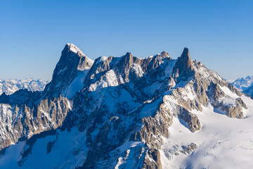 Imposing snow-draped peaks of Aiguille Verte and Les Drus dominate the wintry alpine landscape, their rugged rock faces and sharp ridges highlighted by crisp sunlight beneath a clear blue sky.