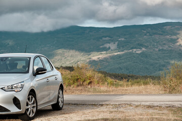 Silver compact car parked on a rural mountain road under cloudy sky, scenic hills in background and copy space. Transportation and travel concept.
