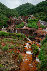 Portrait view of Golden Waterfall near Jiufen, Taiwan, with water flowing over rust-colored mineral rocks and surrounded by lush green vegetation, creating a vibrant vertical nature composition.

