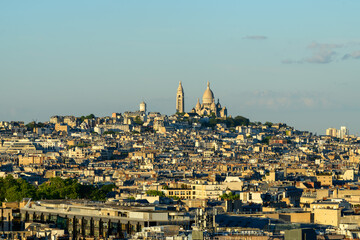 Naklejka premium Sacre-Cur basilica rises above the densely packed rooftops of Montmartre, bathed in warm evening sunlight under a clear blue sky. The elevated view highlights the layered urban landscape and iconic