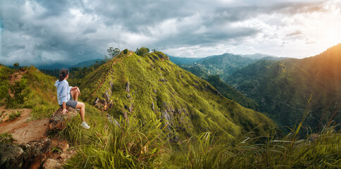 Traveler woman sitting on rock, admiring panoramic sunset view over clouds and green hills from Ella Rock lookout point, Sri Lanka highlands.