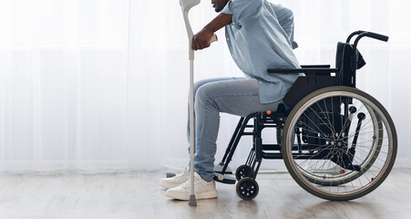 Obraz premium A man using a wheelchair grips his crutches and prepares to stand. He is dressed casually in a light blue shirt and jeans. The room has soft lighting and a bright window.