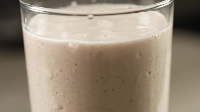 Extreme close-up of a creamy beige smoothie, milkshake, or protein shake in a glass cup, showing thick texture and consistency, representing a healthy breakfast or dessert beverage.