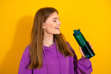 Young woman in purple hoodie smiles while holding a green water bottle on bright yellow background...