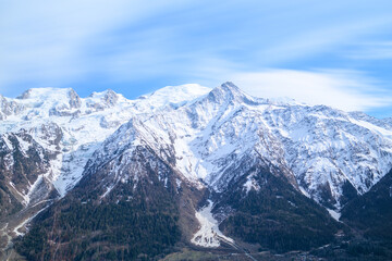 Obraz premium Snow-capped peaks of the Mont Blanc massif tower above dark pine forests and deep valleys, set against a sky streaked with wispy clouds.
