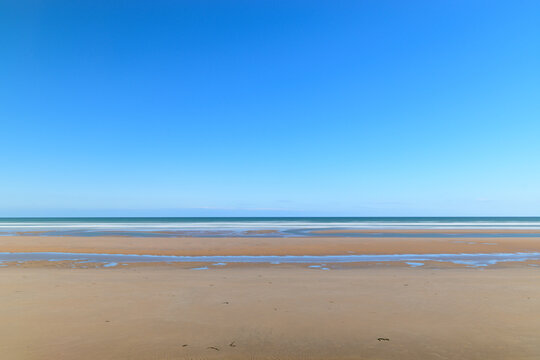 Vast open sands and shallow tidal pools stretch across Omaha Beach beneath a cloudless, vibrant blue sky, creating a peaceful and spacious coastal scene.