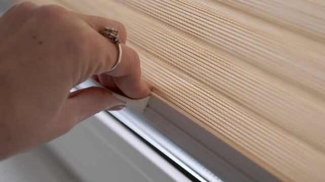 Woman's hand with a ring raises a cordless, pleated cellular window shade using a small tab. Close-up shot follows the motion, highlighting the woven texture of the beige fabric in bright light.