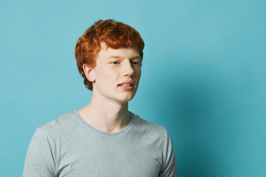 Young man with red hair wearing gray t-shirt looking aside with neutral expression on blue background. Portrait of caucasian male with casual outfit and short hairstyle.