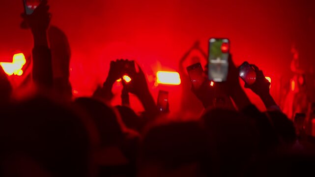 Silhouetted crowd at a live music concert. Fans raise hands and record the performance on smartphones under intense, vibrant red stage lighting. Energetic and exciting atmosphere.