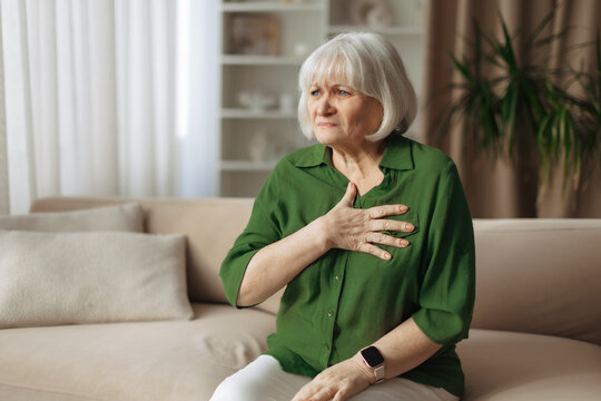 Elderly caucasian woman holding chest while sitting on sofa at home, senior female experiencing discomfort or anxiety, health concern and retirement lifestyle concept indoors