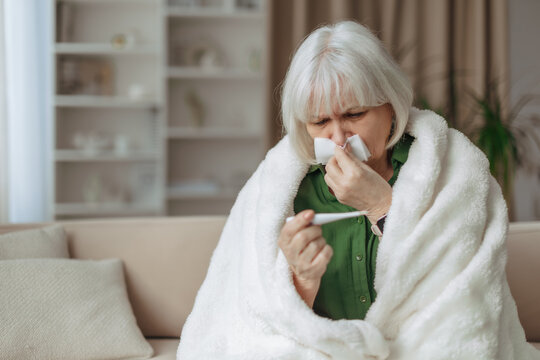 Senior caucasian woman wrapped in blanket holding thermometer at home, elderly female checking body temperature, sickness and home care retirement lifestyle concept