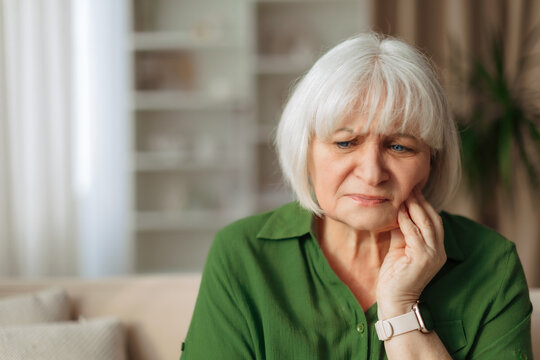 Elderly caucasian woman touching cheek with worried expression at home, senior female feeling tooth or facial pain, retirement lifestyle and healthcare awareness concept