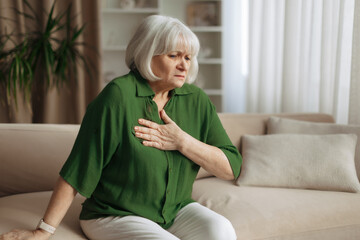 Senior caucasian woman leaning forward and touching chest at home, elderly female feeling shortness...