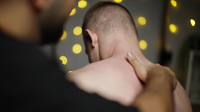 Close-up therapeutic shoulder and neck massage on a bare back, deep tissue pressure easing muscle tension and soreness, with soft bokeh lights creating a calm wellness atmosphere