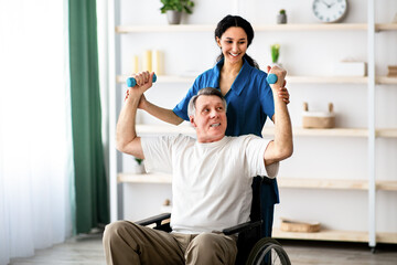 A man using a wheelchair lifts weights with guidance from a healthcare worker in a well-lit therapy...