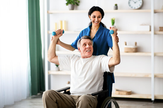 A man using a wheelchair lifts weights with guidance from a healthcare worker in a well-lit therapy space. The setting includes shelves with various items, creating a supportive environment.