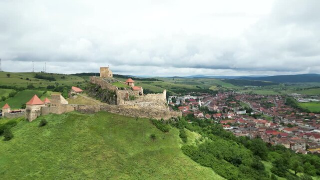 Aerial drone view of Rupea Fortress overlooking Rupea village, Romania, set on a green hill with historic medieval walls, dramatic clouds and a scenic Transylvanian landscape.