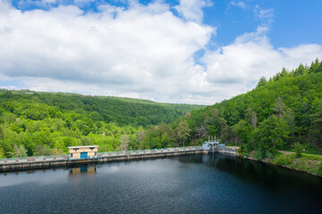 Fototapeta premium A small utility building stands on a concrete dam spanning a calm lake, surrounded by dense green forest and rolling hills beneath a partly cloudy blue sky at Lac de Chaumecon.