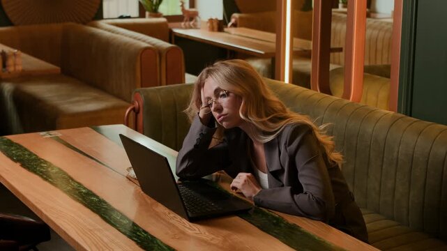 Tired young blonde woman in a blazer and glasses leans on her hand and looks at a laptop in a stylish cafe lounge. Slow motion lateral camera movement highlights work stress, fatigue, and burnout.