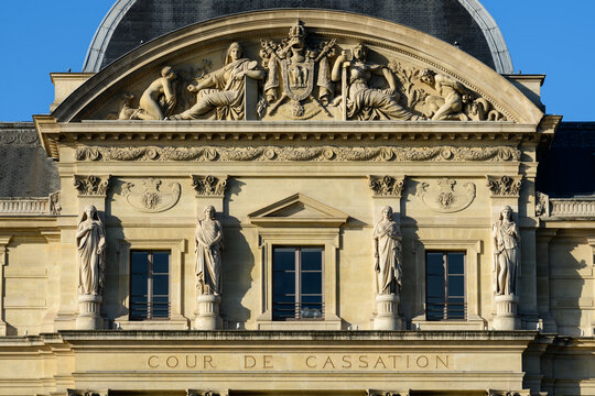 Detailed view of classical stone statues and ornate reliefs adorning the pediment and facade of the Cour de Cassation in Paris, illuminated by crisp sunlight against a vivid blue sky.