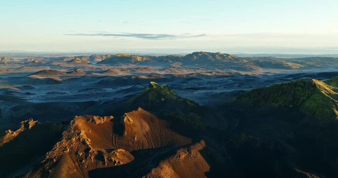 Drone glides over Iceland highlands at dawn, with blue haze on volcanic plains, a mossy peak and ochre slopes catching first light, long shadows, and a quiet endless horizon.