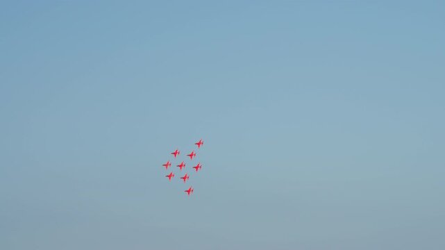 A squadron of nine red jets performs a synchronized maneuver in a clear blue sky. They fly in a tight diamond formation, leaving long white smoke trails behind them as they fly away.