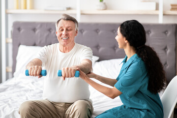 Fototapeta premium A senior man with gray hair smiles as he lifts light dumbbells in a bright room. A woman with dark hair supports him, encouraging his exercise routine. This scene shows care and physical activity.