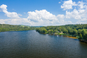 Fototapeta premium Bright summer scene at Lac des Settons featuring calm blue water, a tree-lined shoreline, and boats moored near the edge beneath a sky filled with fluffy clouds.