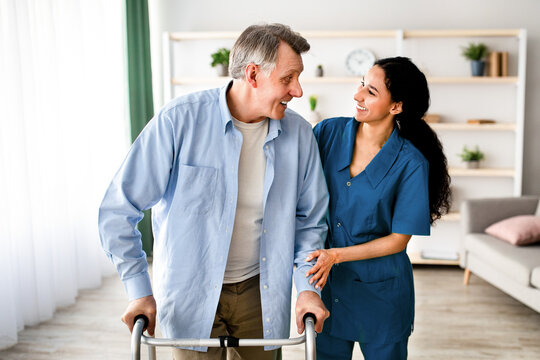 A senior man is using a walker with assistance from a caregiver. They are both smiling in a well-lit room that has light-colored furniture. This moment shows warmth and care.