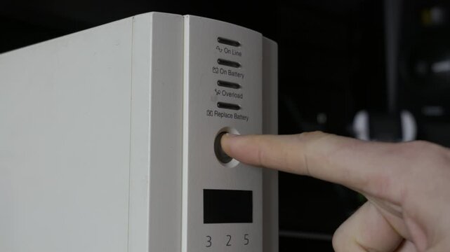 A male finger presses the circular power button on a white UPS battery backup unit, activating the LED status lights. Concept of electrical safety, surge protection, and data security.