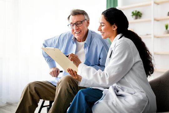 A woman in a white coat shares information on a clipboard with an older man in glasses. They are seated in a well-lit clinic. The man is smiling and engaged in the conversation.