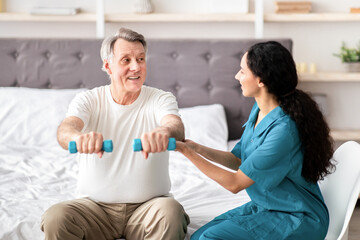 Obraz premium A senior man is sitting on a bed holding small weights as a woman provides encouragement and assistance. They are in a bright room designed for physical activity.