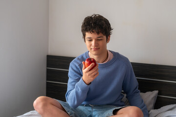 Handsome young man in a blue sweater and denim shorts sitting on the bed with an apple in his hands, concept of healthy eating and teenage diet