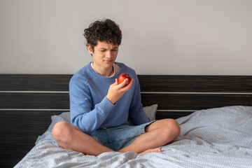 A handsome young man in a blue sweater and denim shorts sits on a bed with an apple in his hands and looks at it with displeasure, the concept of healthy eating and teenage choice