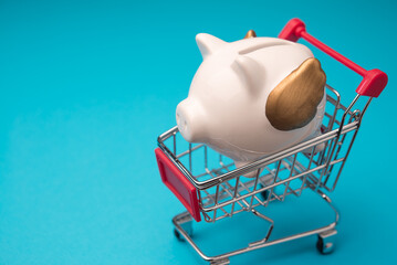 White piggy bank in shopping trolley on blue background copy space. Savings and financial management planning for shopping, expenses, cost of living, financial literacy, save money and so on concept.