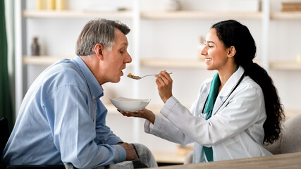 Obraz premium A woman with dark skin is feeding a senior man with gray hair in a well-lit room. They are both smiling and engaged in a pleasant moment during a meal.