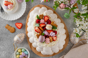 Obraz premium Easter egg-shaped Pavlova cake with whipped cream, topped with fresh berries and assorted sweets on a wooden board on a concrete background. Top view, copy space.